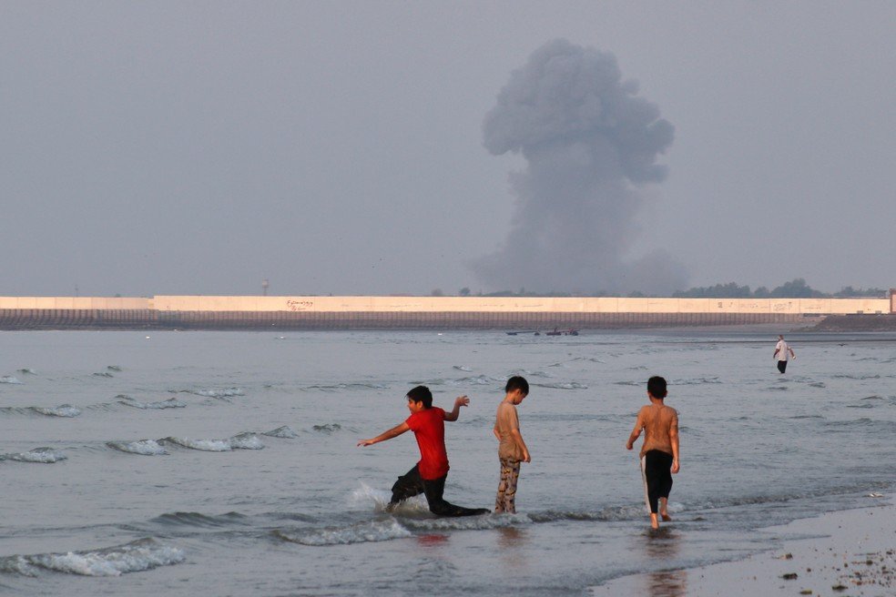 Três meninos brincam nas águas rasas do Estreito de Ormuz, enquanto uma coluna de fumaça sobe de uma explosão ao fundo, perto de Bandar Abbas, Irã.